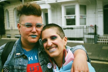 Photo credit: Matthew Jacobs Morgan, taken at Trans Pride 2019 Photo of Al (left) and Amelia (right). Al is a round white human with fantastically curly brown hair with shaved sides, wearing clear rimmed glasses and an excellent denim jacket with a nonbinary flag pin on the collar. Amelia is a slim white human with buzzed brown hair wearing very long colourful earrings and with a trans flag wrapped around their shoulders like a superhero cape. Both have sparkly blue, white and pink makeup on. Al has their arm casually around Amelia’s shoulders and Amelia smiles at the camera. 